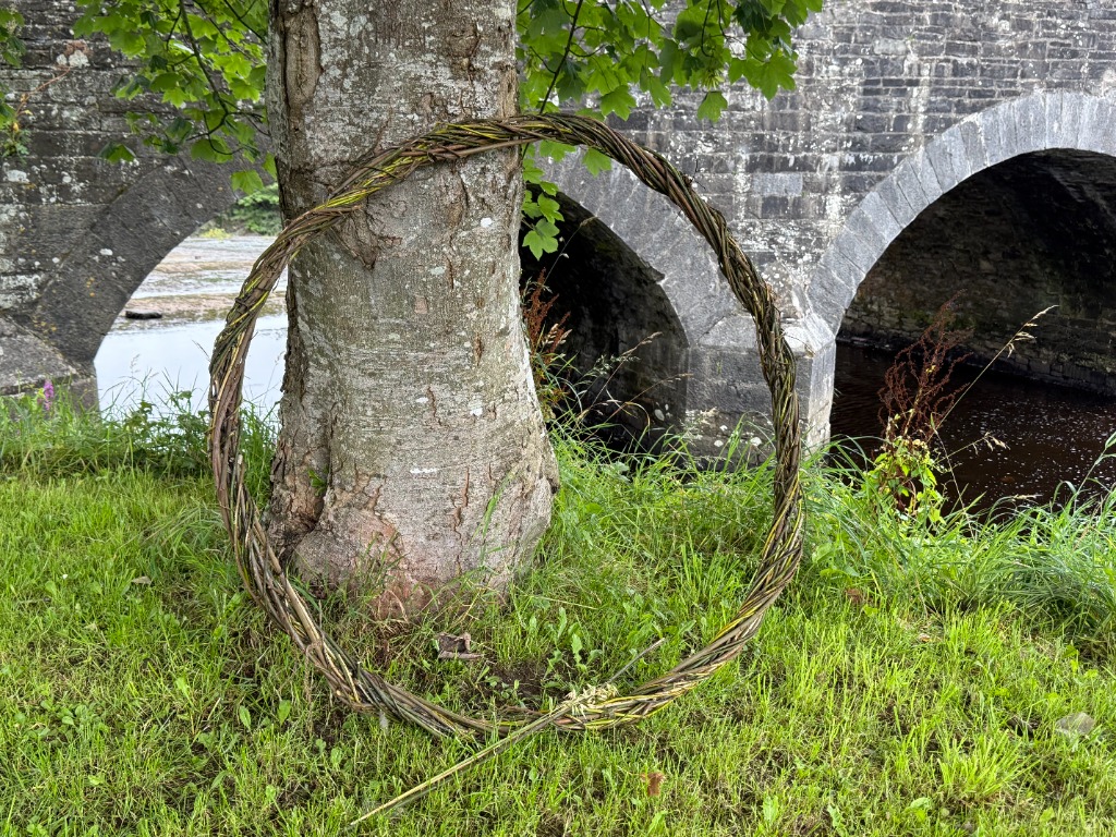 Un aro tejido de sauce apoyado contra un árbol junto a un antiguo puente de piedra